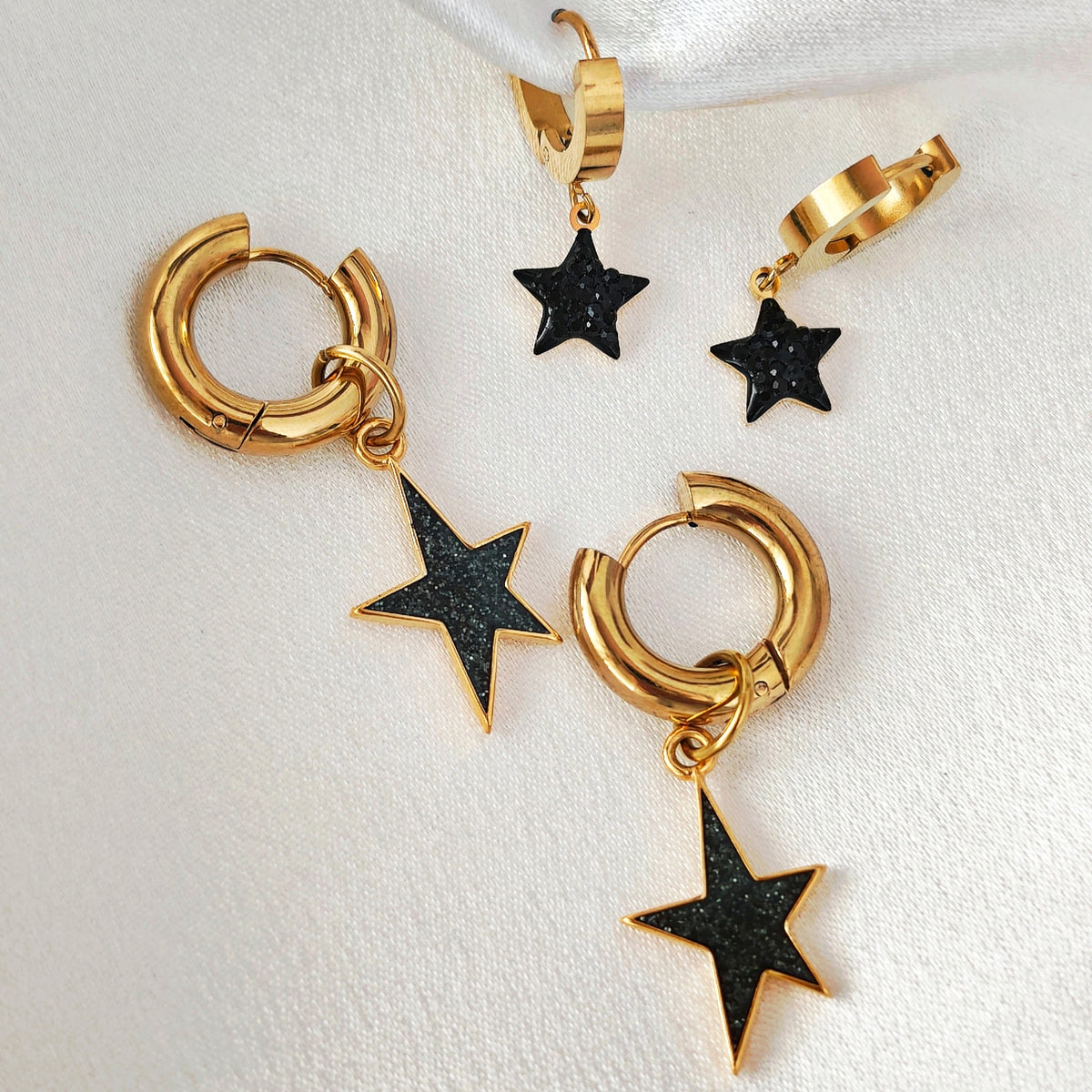 Small and large gold-tone hoop earrings with black glitter star charms on white satin.