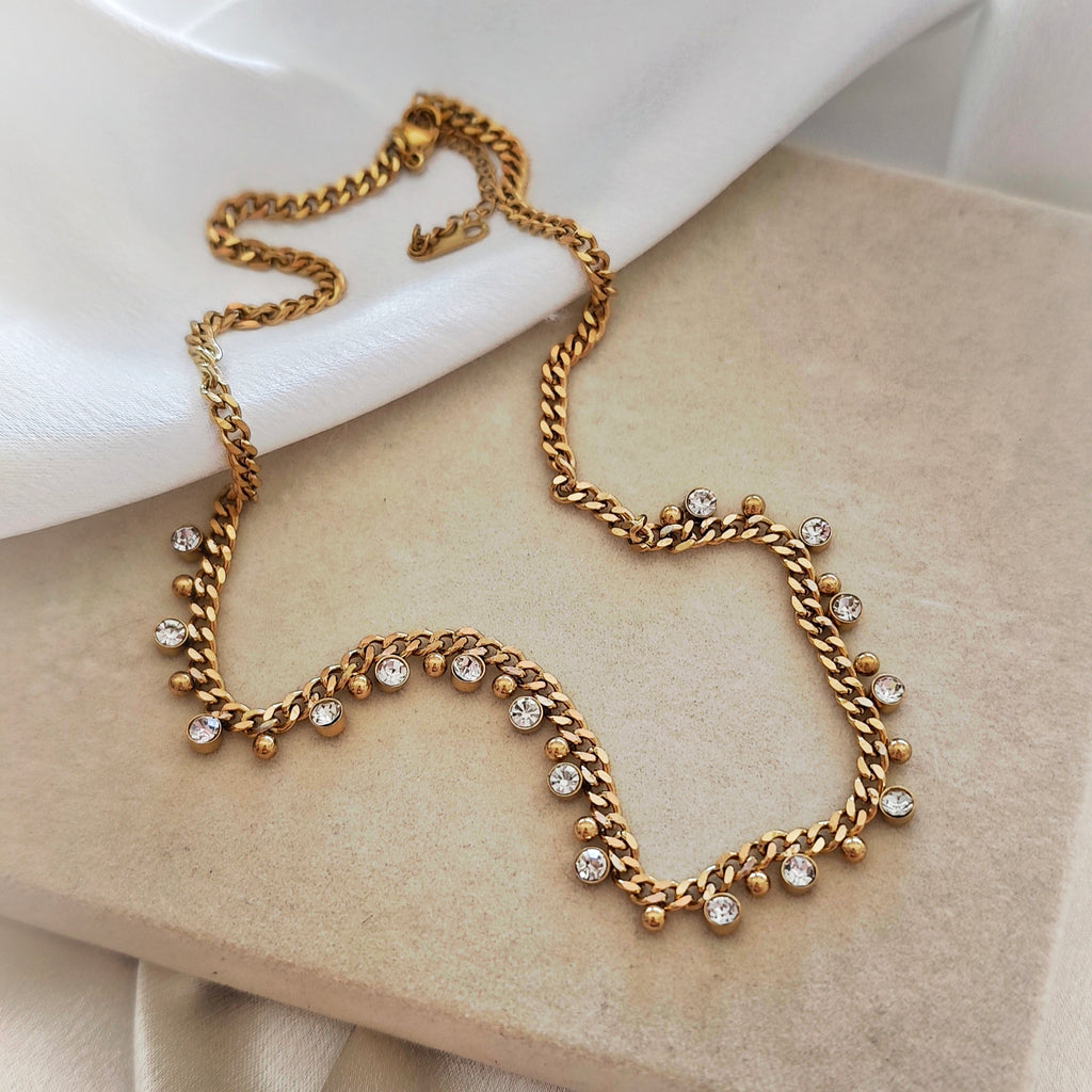 Gold-tone steel necklace with dangling clear stones and small gold-tone beads on a beige surface.