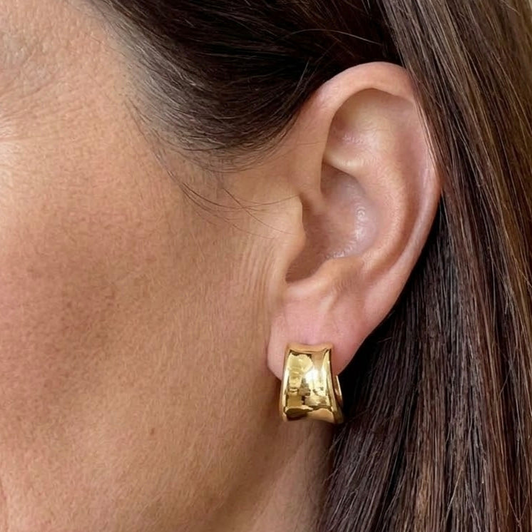 Polished gold-tone wide curved steel hoop earring worn on an ear.