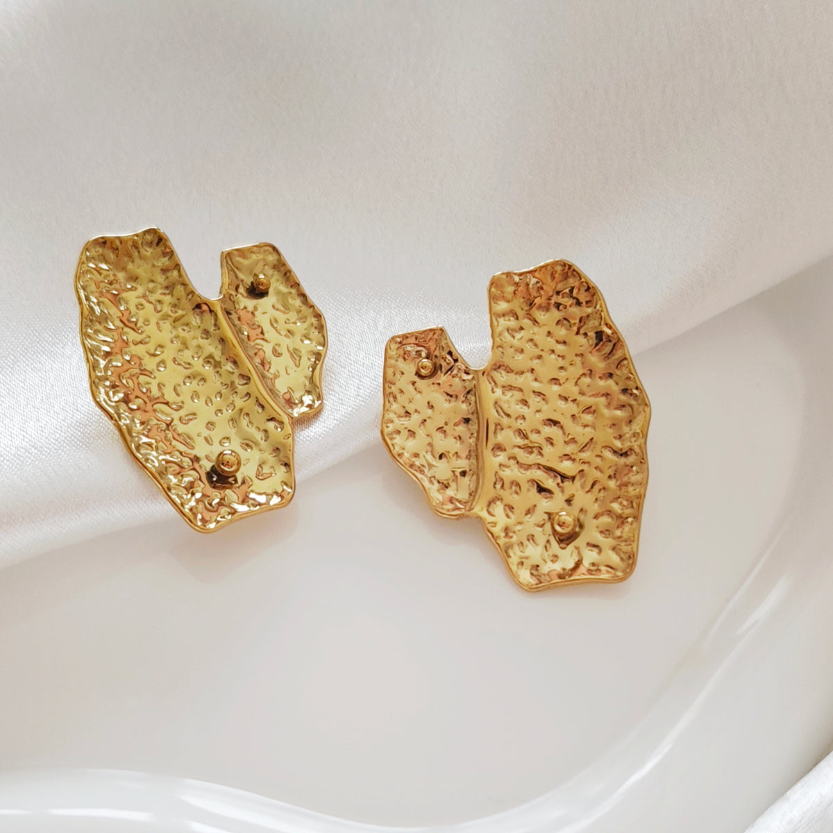 Pair of gold-tone earrings with irregular folded shapes and textured surfaces on ivory satin.