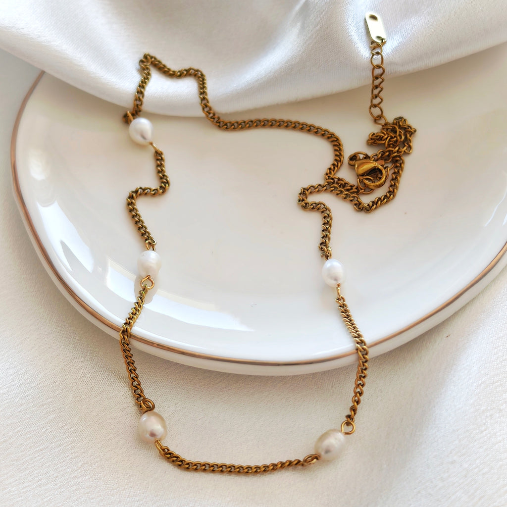 Gold-tone steel necklace with white beads displayed on a white plate.