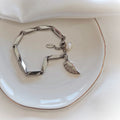 Silver-tone steel bracelet with geometric links, a wing charm and a white bead on a plate.