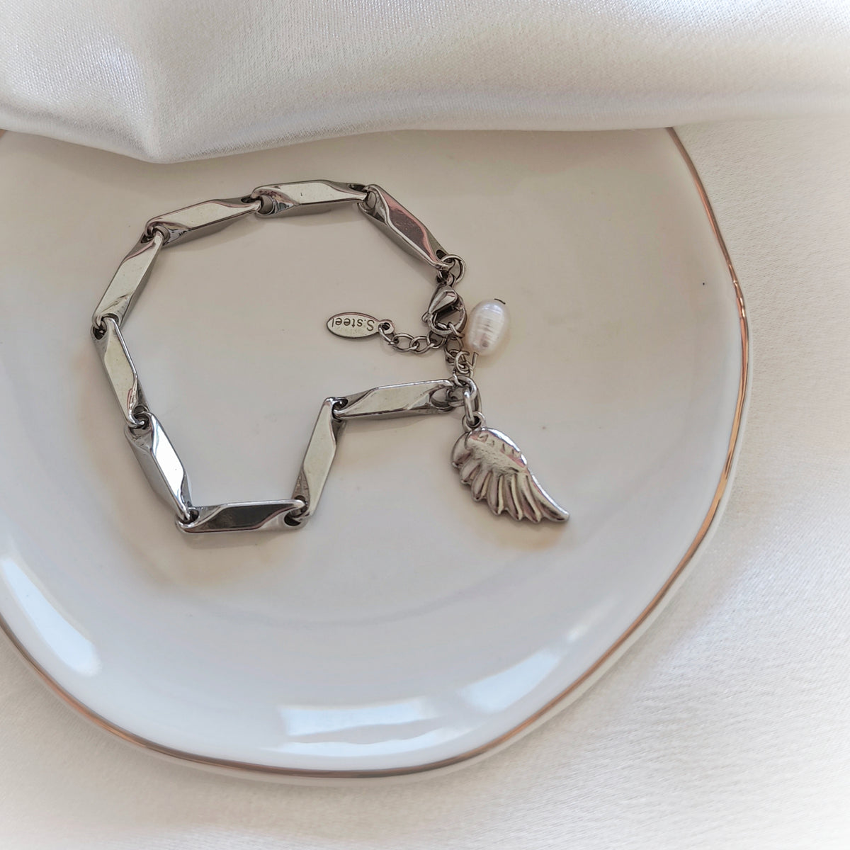 Silver-tone steel bracelet with geometric links, a wing charm and a white bead on a plate.