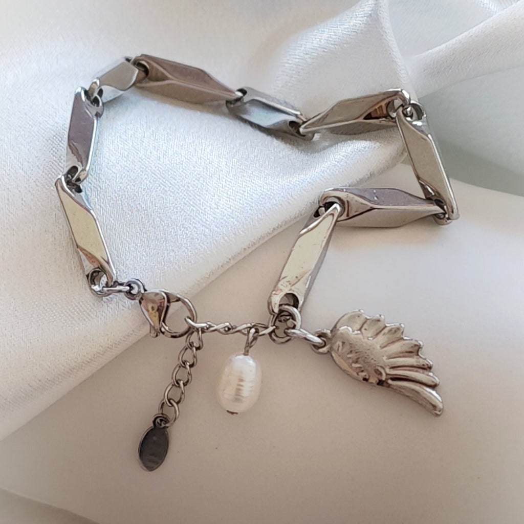 Close-up of silver-tone geometric link bracelet with wing charm and white bead on ivory satin.