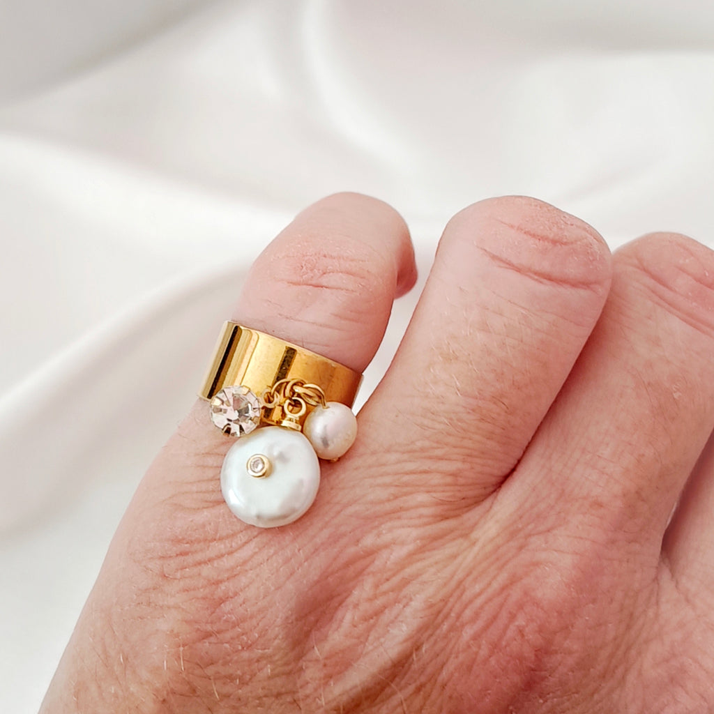 Gold-tone steel ring with dangling pearls and a clear decorative stone worn on a hand.
