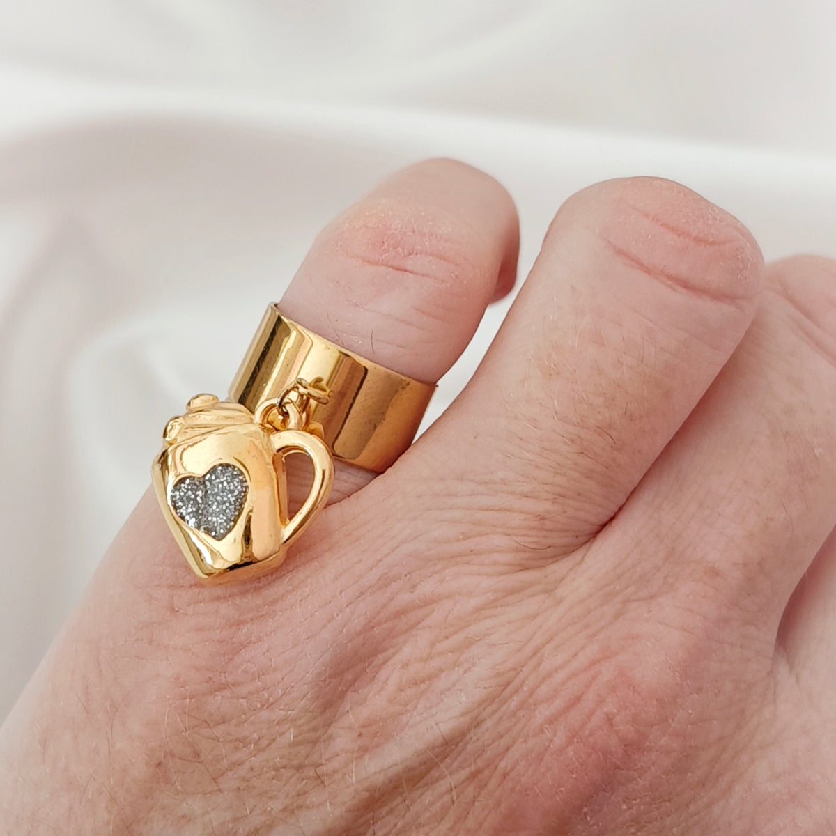 Gold-tone open band ring with a dangling mug charm and sparkling heart on ivory fabric.