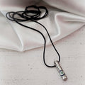 Silver-tone cylindrical steel pendant with an eye detail and black cord on ivory fabric.