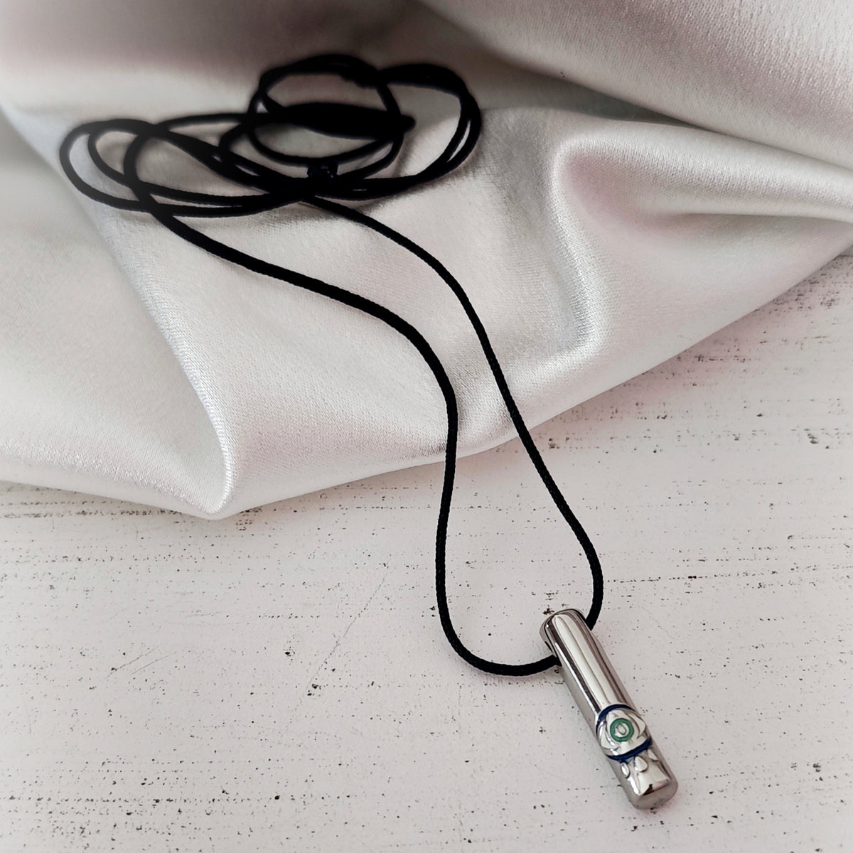 Silver-tone cylindrical steel pendant with an eye detail and black cord on ivory fabric.
