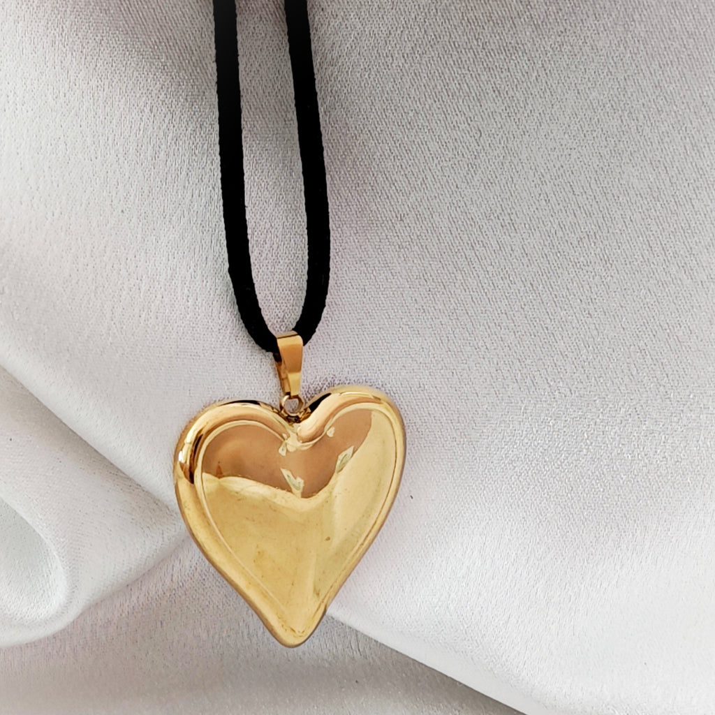Polished gold-tone steel heart pendant on black suede cord against ivory fabric.