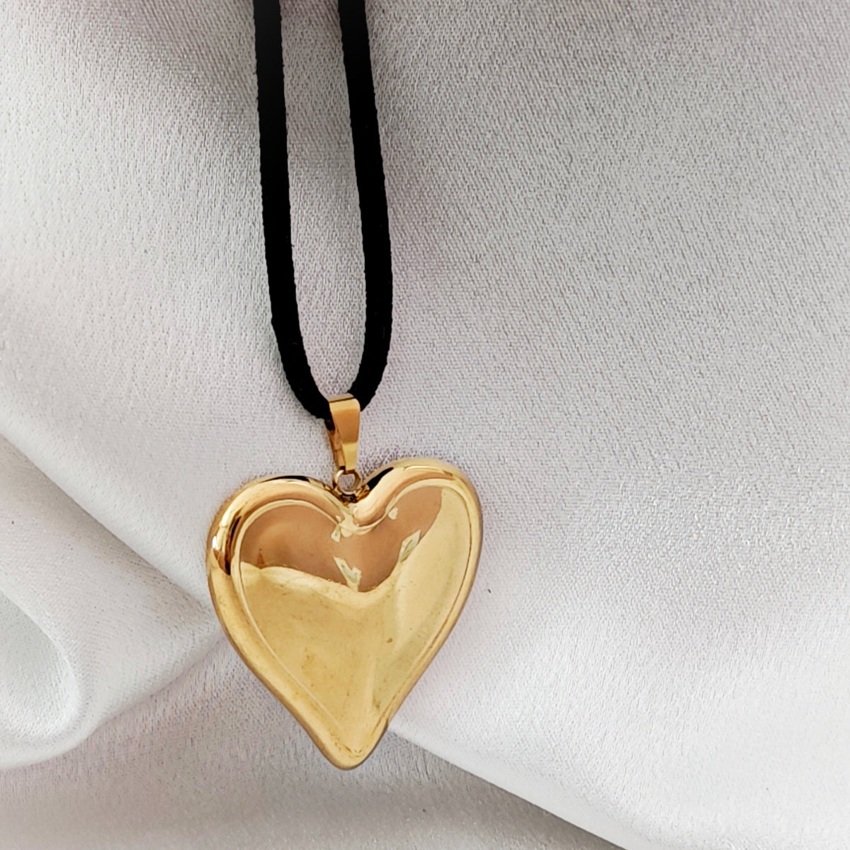 Polished gold-tone steel heart pendant on black suede cord against ivory fabric.