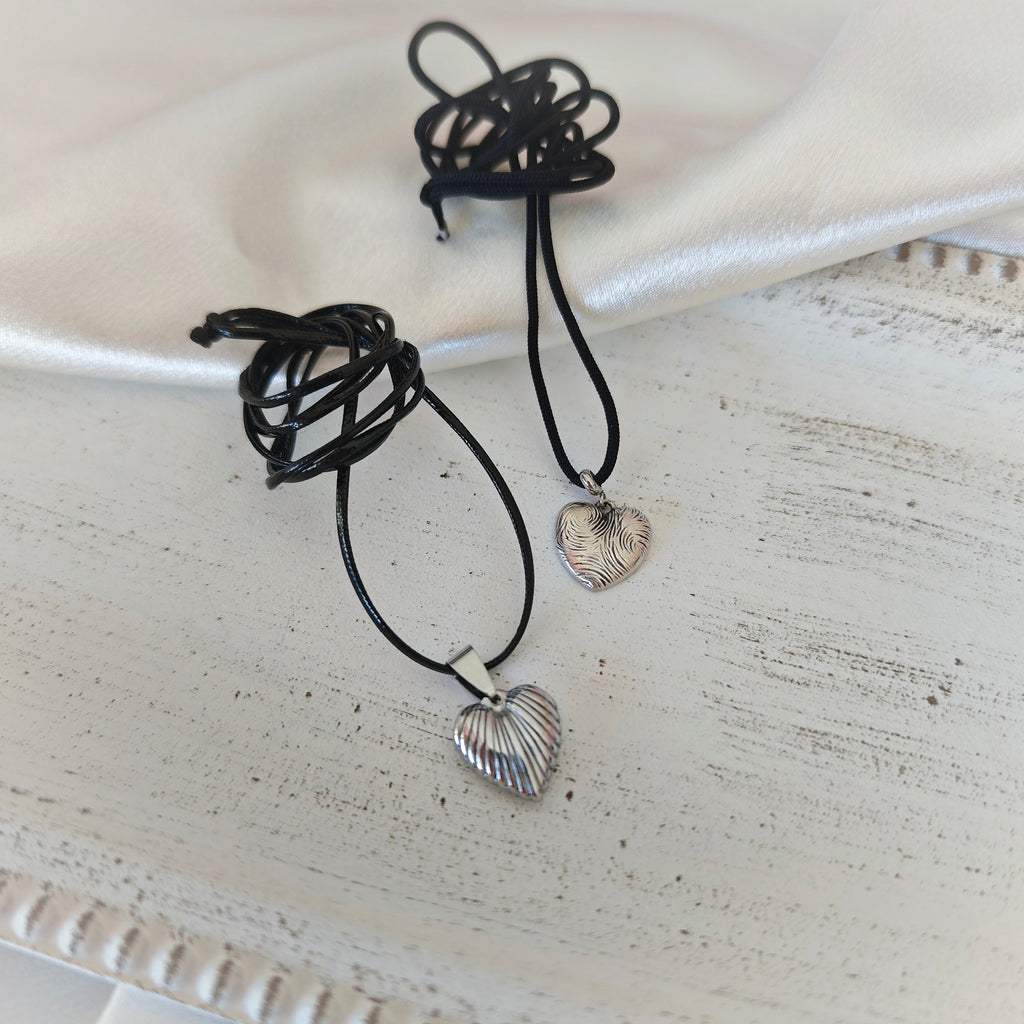 Two heart pendant necklaces with black leather cords, one ribbed stainless steel heart and one silver-plated heart with wavy engravings