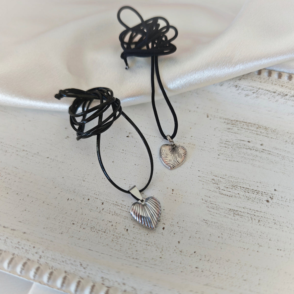 Two heart pendant necklaces with black leather cords, one ribbed stainless steel heart and one silver-plated heart with wavy engravings
