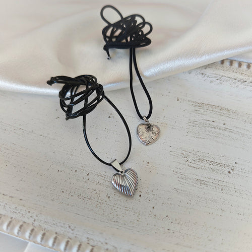 Two heart pendant necklaces with black leather cords, one ribbed stainless steel heart and one silver-plated heart with wavy engravings
