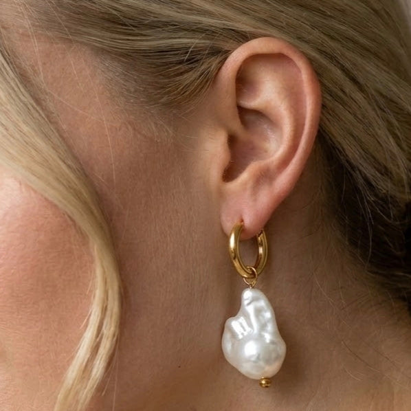 Gold-tone hoop earring with an irregular white pearl drop worn on an ear