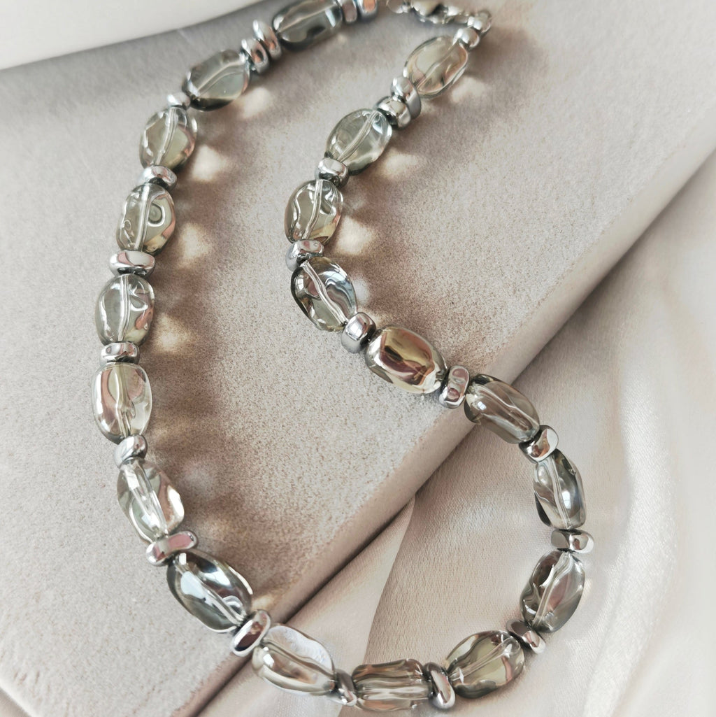 Lucent necklace with translucent grey oval crystal beads and silver-tone hematite details on a beige display.