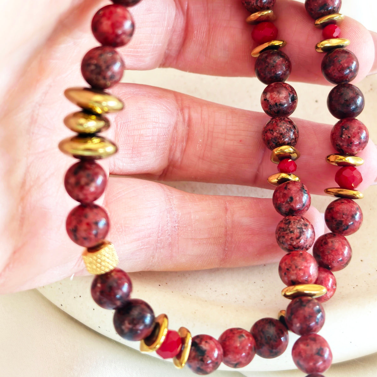 Burgundy semiprecious stone necklace with dark mottled beads and gold-tone details held in a hand.